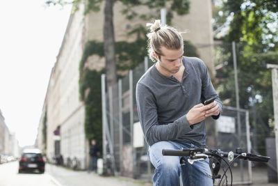 Young man text messaging through mobile phone while sitting on bicycle at street