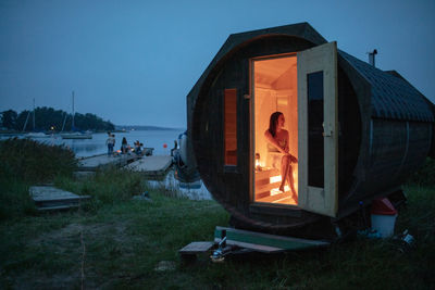 Full length of mid adult woman sitting in sauna cabin by sea against sky