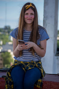 Portrait of smiling young woman using mobile phone while sitting on retaining wall