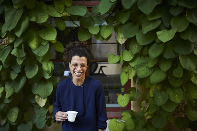 Happy young female architect holding coffee cup against creeper plants in backyard