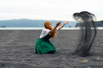 Side view of woman playing with sand at beach against sky