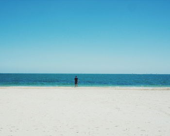 Distant view of man standing at sea shore against blue sky