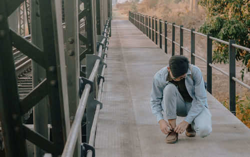 Full length of young man tying shoelace on bridge