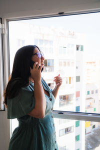 Brunette spanish girl dressed on a business serius look talking on the phone at the window from her apartment during the afternoon in palma de mallorca, spain during coronavirus confinement