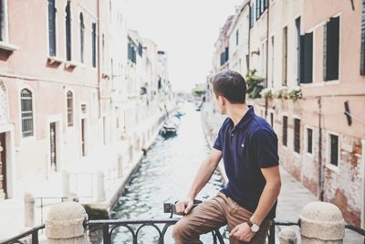 Man looking away while sitting on railing over canal amidst buildings