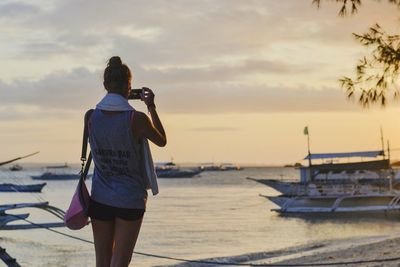 Rear view of woman photographing boats moored in sea at sunset