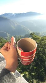 Midsection of person holding tea cup against mountains