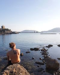 Rear view of shirtless man sitting on rock by lake against sky