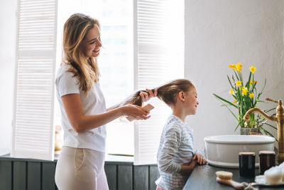 Young mother woman with little tween girl daughter in pajamas combs her hair in bathroom at home