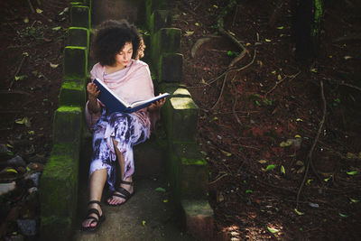 Beautiful young woman with a curly hair sitting on a cement staircase with moss reading a book