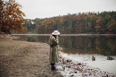 Side view of woman wearing hat photographing birds while standing by lake against sky
