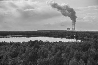 Smoke emitting from factory by lake against sky