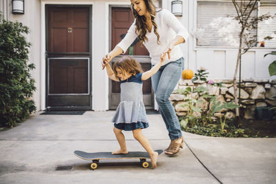 Smiling daughter balancing over skateboard with help of mother on footpath