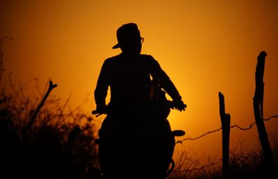 Silhouette man riding motor scooter against orange sky