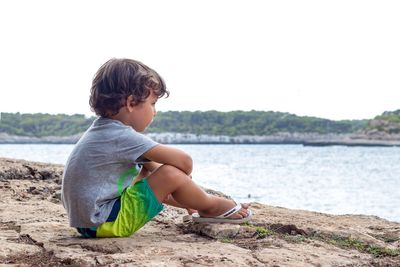 Rear view of boy sitting on beach against clear sky