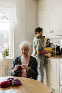 Senior woman knitting while female caregiver working in kitchen