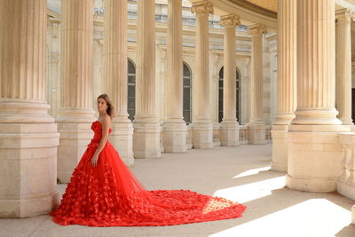 Portrait of woman wearing red gown standing by architectural columns