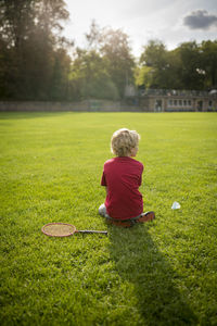 Rear view of boy with racket and shuttlecock sitting on grassy field against trees