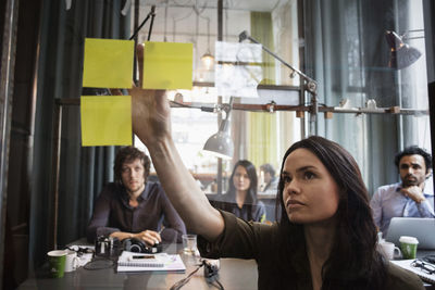 Creative business people looking at female colleague sticking adhesive note on window seen through glass