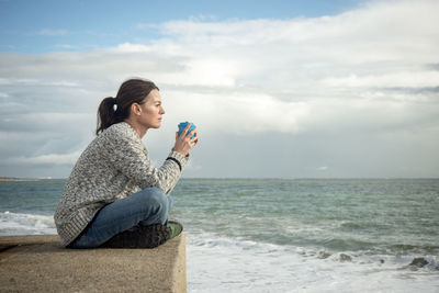 Side view of young woman sitting at beach against sky
