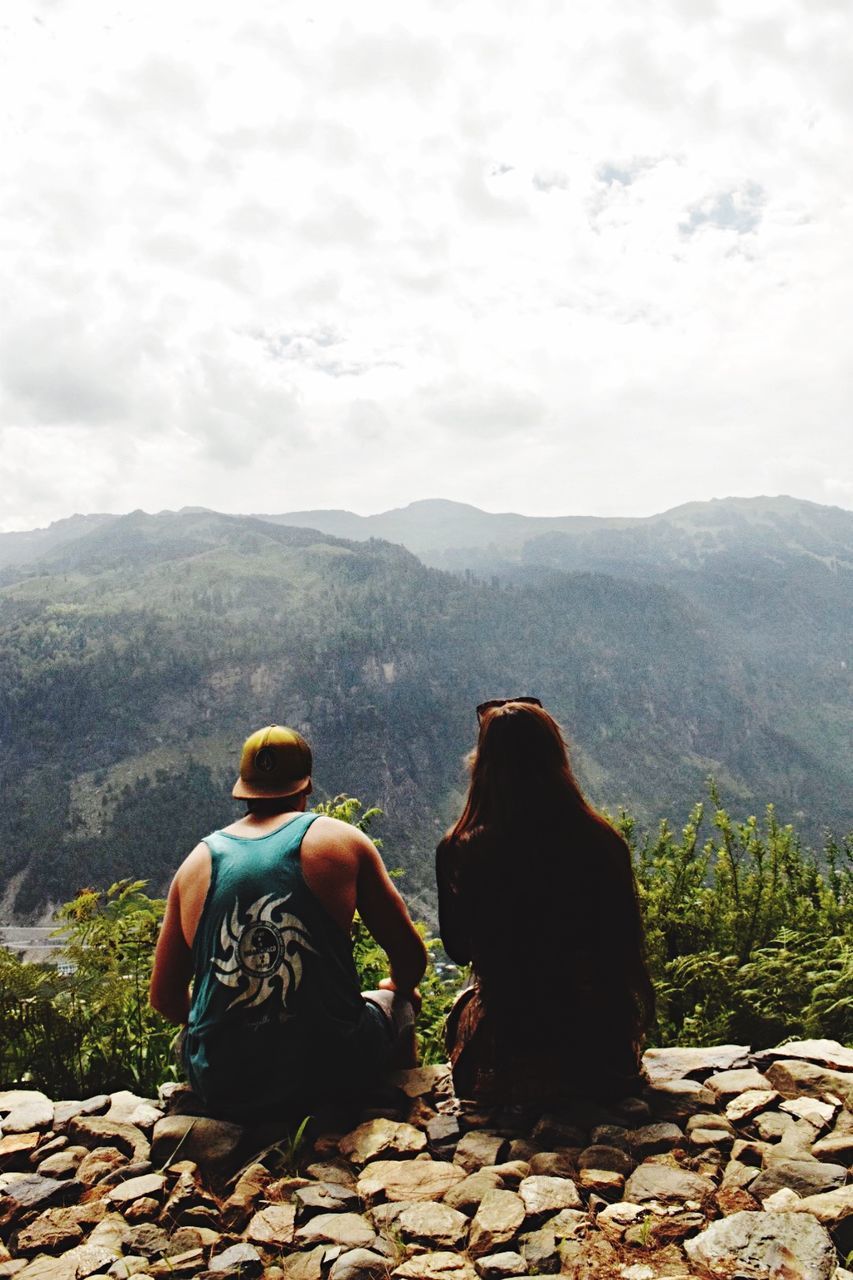 REAR VIEW OF FRIENDS SITTING ON ROCKS AGAINST MOUNTAIN