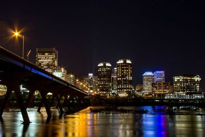 Illuminated bridge over river by buildings against sky at night