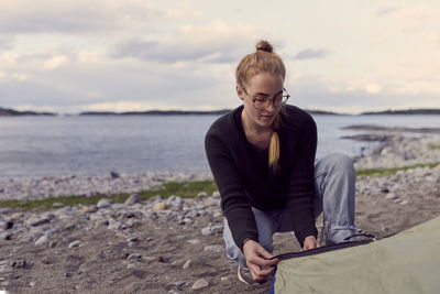 Young woman crouching while setting up tent at beach against sky