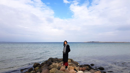 Portrait of woman standing on rocks at beach against cloudy sky