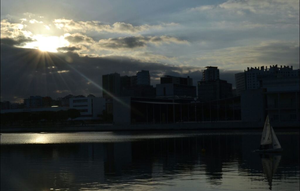 architecture, building exterior, water, built structure, sun, sunbeam, sky, city, reflection, sunlight, waterfront, lens flare, river, sunset, cloud - sky, skyscraper, cloud, cityscape, outdoors, no people