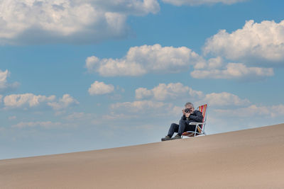 View of an adult male sitting on a beach chair at the top of a dune taking a picture