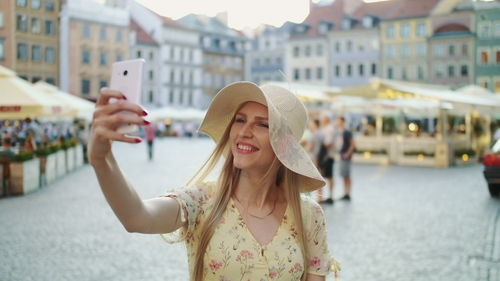 Cheerful young woman wearing hat taking selfie while standing on street against buildings