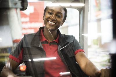 Portrait of happy female warehouse worker sitting in forklift seen through windshield