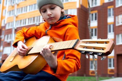 Portrait of caucasian blond boy in orange jacket playing acoustic guitar outdoors on autumn day