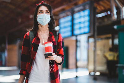 Low angel view of young woman wearing mask holding coffee cup standing at airport