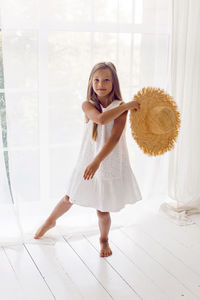 Girl in a white dress standing next to the window of the house and holding a straw hat