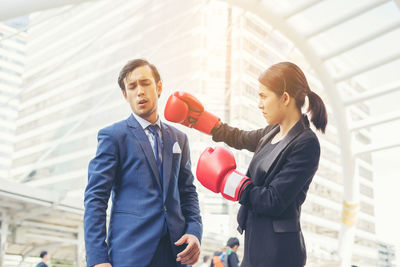 Young businesswoman hitting colleague with red boxing gloves against buildings in city