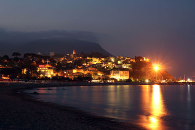 Illuminated buildings by sea against sky at night