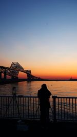 Silhouette person standing by tripod against bridge during sunset