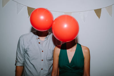 Couple face covered with red balloons against bunting flag on wall