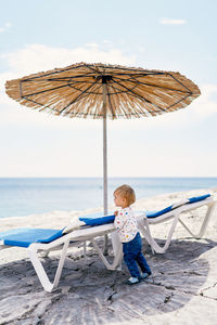Boy sitting on chair at beach against sky