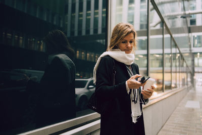 Businesswoman with coffee using smart phone while standing on sidewalk against modern building in city