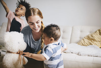 Smiling mother showing teddy bear to toddler while boy playing by sofa at home