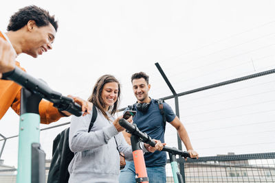 Low angle view of friends using mobile phone while holding push scooters in city against sky