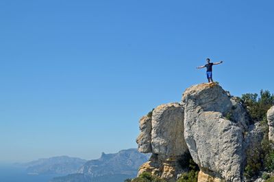 Young man with arms outstretched standing on boulder against clear blue sky