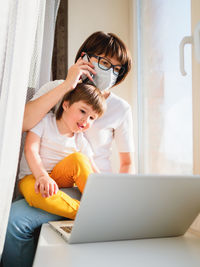 Woman in medical mask remote works from home. she sits on window with laptop and cute toddler boy