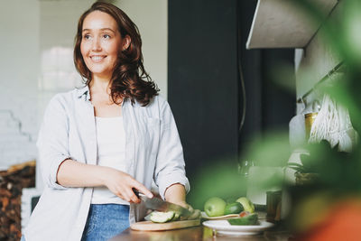 Smiling woman looking away while cutting fruits at kitchen