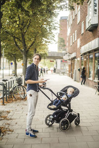 Side view of smiling man looking way while standing by son on baby stroller in city