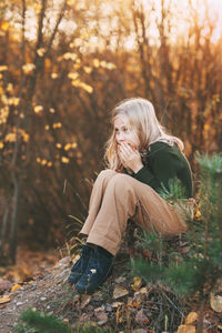 A beautiful teenage girl with blond hair sits thoughtfully in an autumn park, warms her hands