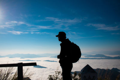 Side view of silhouette backpacker wearing cap while standing on mountain against sky