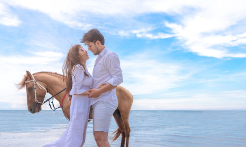 Side view of couple standing by horse on beach against sky